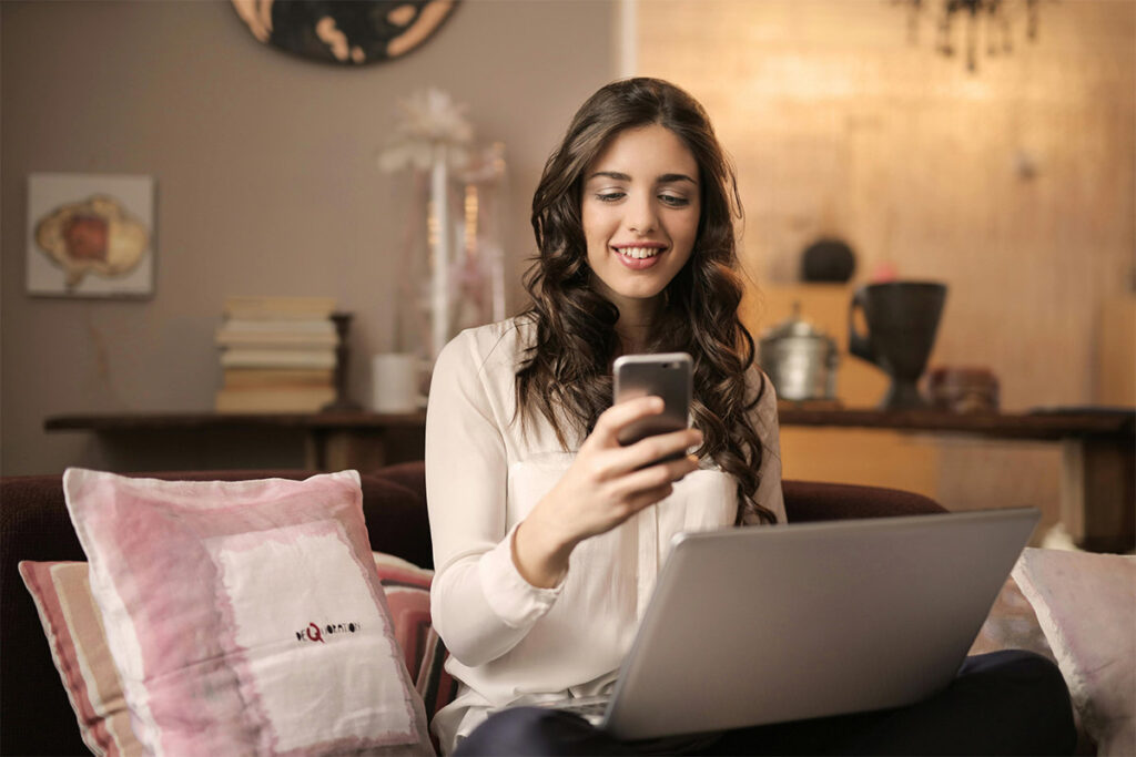 A smiling woman using her smartphone while working on a laptop in a cozy home setting.