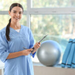 Smiling physical therapist in blue scrubs holding a clipboard in a modern clinic setting with exercise equipment in the background.