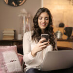 A smiling woman using her smartphone while working on a laptop in a cozy home setting.