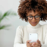 Smiling woman wearing glasses and a white sweater using her smartphone, representing digital communication and email marketing.