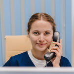 Friendly female medical receptionist in navy scrubs answering a phone call at the front desk, with a professional and welcoming smile.