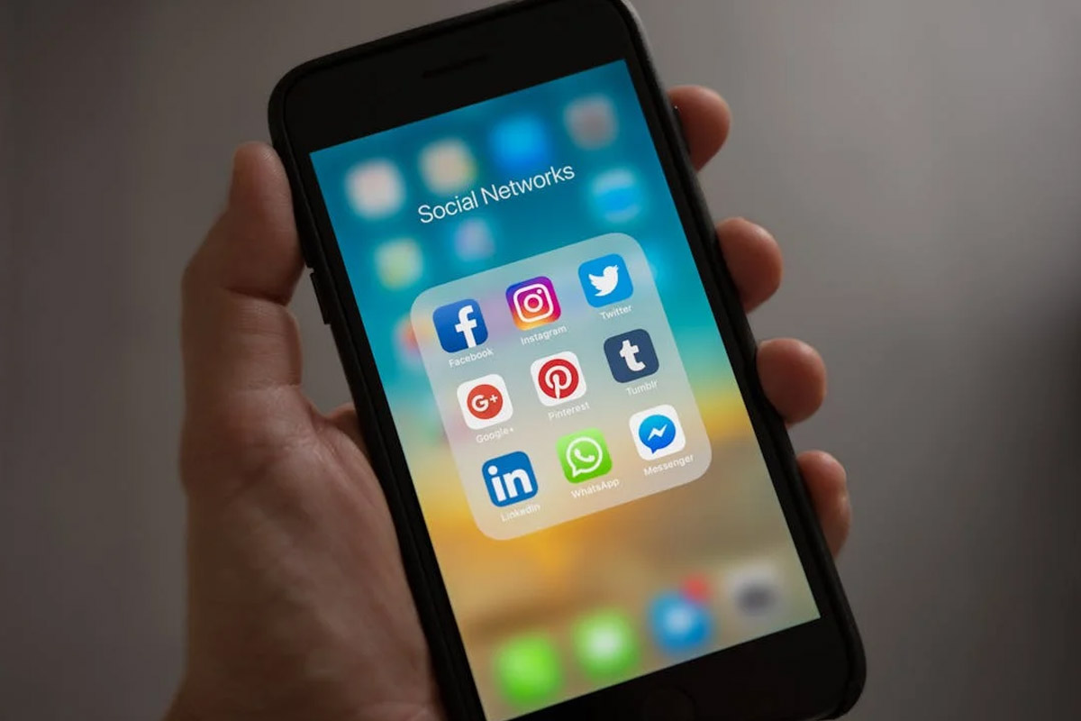 A person holding a smartphone displaying a folder labeled "Social Networks" with various social media app icons, including Facebook, Instagram, Twitter, and LinkedIn.